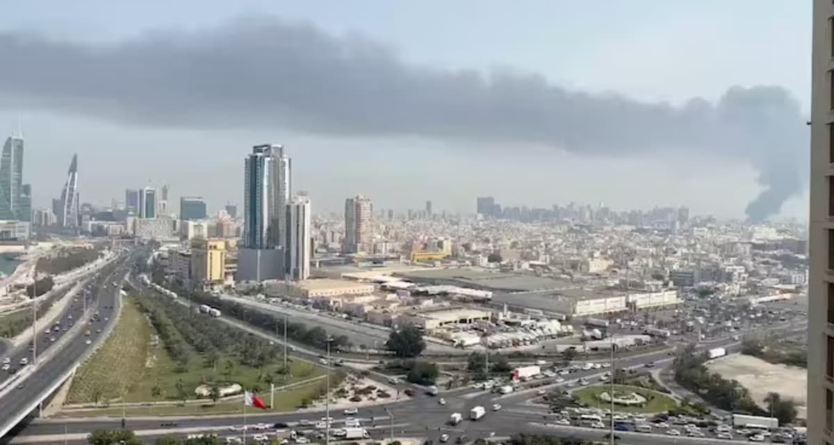 Aerial cityscape of Bahrain showing modern high-rise buildings, highways, industrial areas, and dense urban development under with smoke from an Iranian attack in the background.